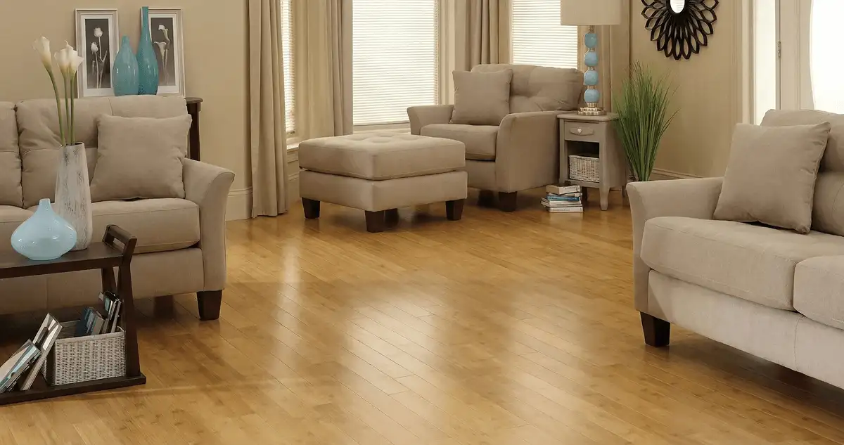 A living room with professionally installed bamboo flooring, beige sofas, a matching ottoman, a side table with a blue lamp, vases, framed pictures, and large windows with blinds and curtains in Baton Rouge, LA