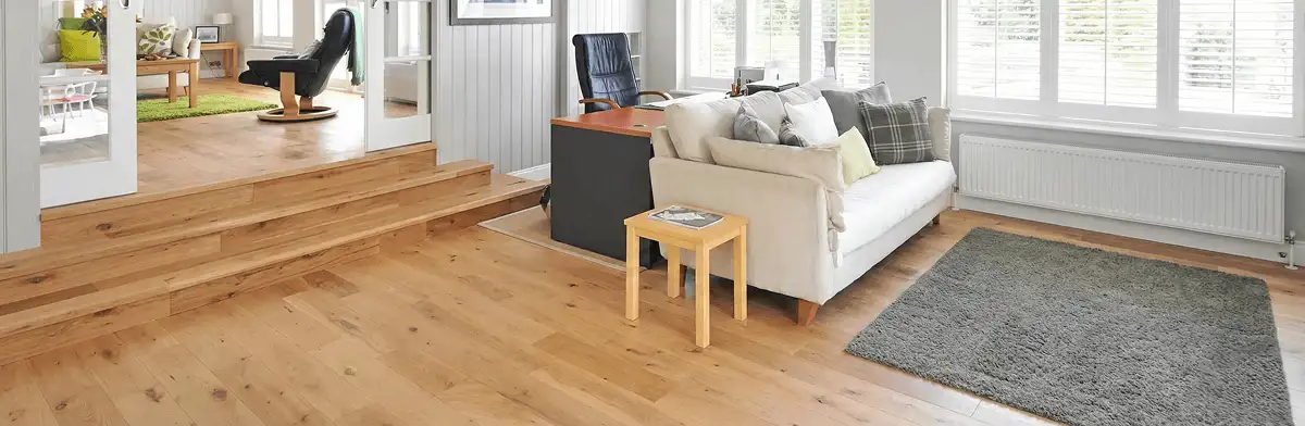 A modern living room with professionally installed laminate flooring, a beige sofa, a small side table, a gray rug, and large windows with white shutters in Baton Rouge, LA